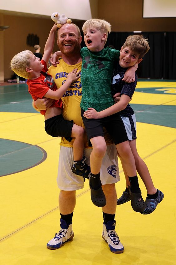 The Ricotta family: a Ken Chertow wrestling camp coach who brings his own kids, laughing on the mat with boys in his arms