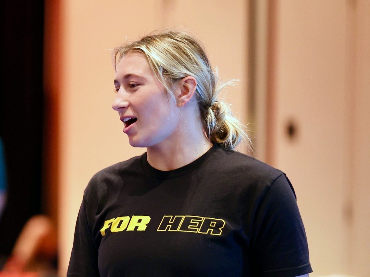 A coach in a black FOR HER shirt talking to campers at Ken Chertow's girls wrestling camp