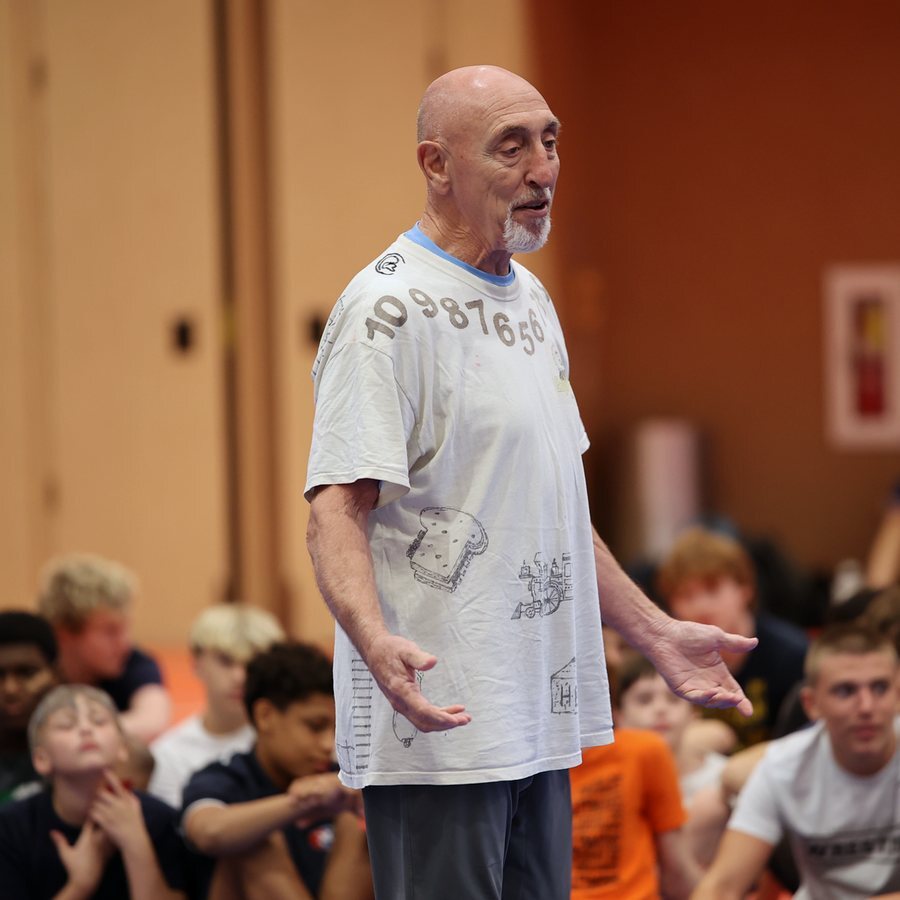 Wade Schalles standing and talking to campers seated on the mat at Ken Chertow's summer wrestling camp