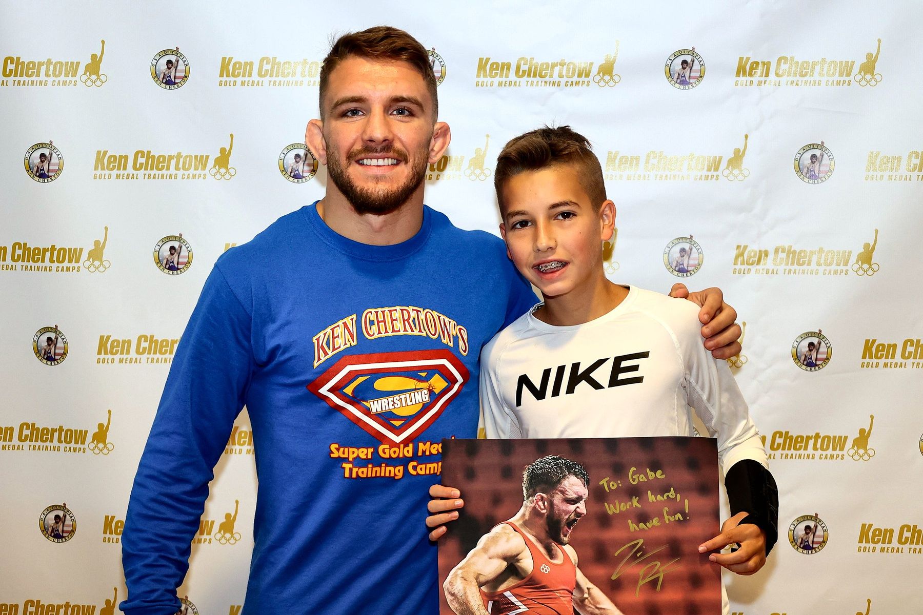 A staff member posing with a camper in front of the Ken Chertow backdrop at summer wrestling camp