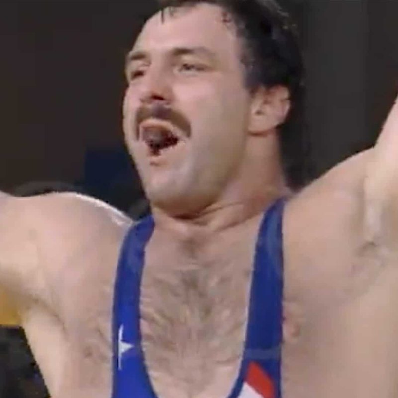 Bruce Baumgartner raising both arms in celebration, in a USA singlet, Ken Chertow wrestling camp coach