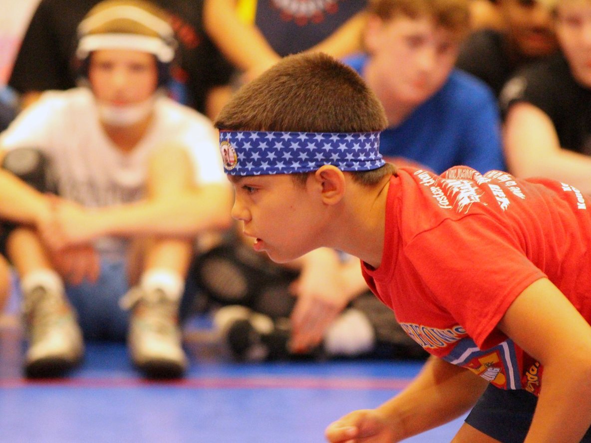 A young wrestler in a stars headband down in his stance at youth wrestling camp