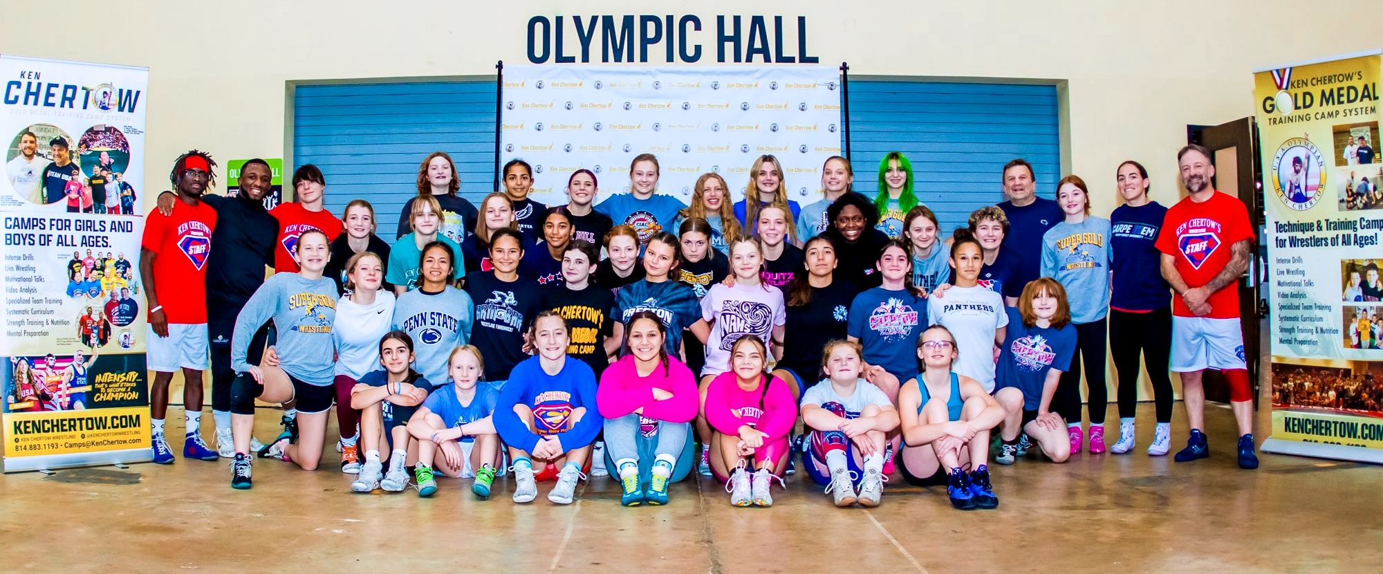Girls and coaches at a past SuperGirl wrestling camp, posed between two Ken Chertow camp banners