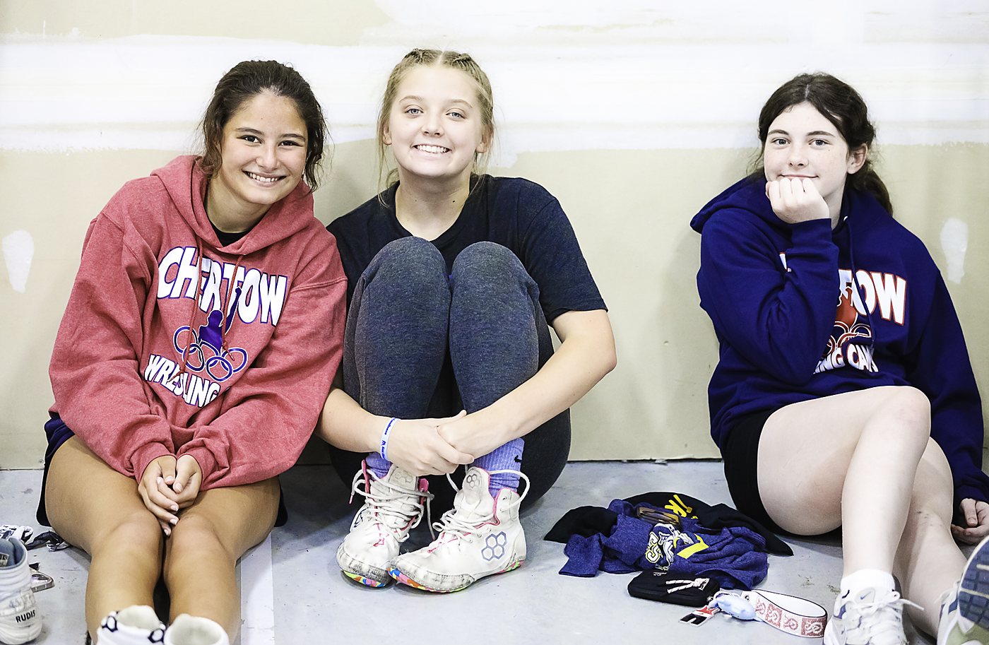 Three girls sitting together against the wall and smiling, one in a red Chertow hoodie