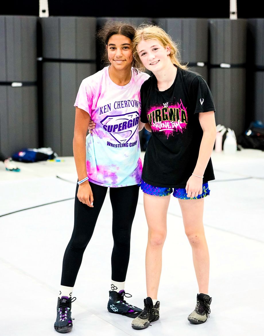 Two girls smiling arm in arm on the mat at SuperGirl summer wrestling camp, one in a tie-dye camp shirt